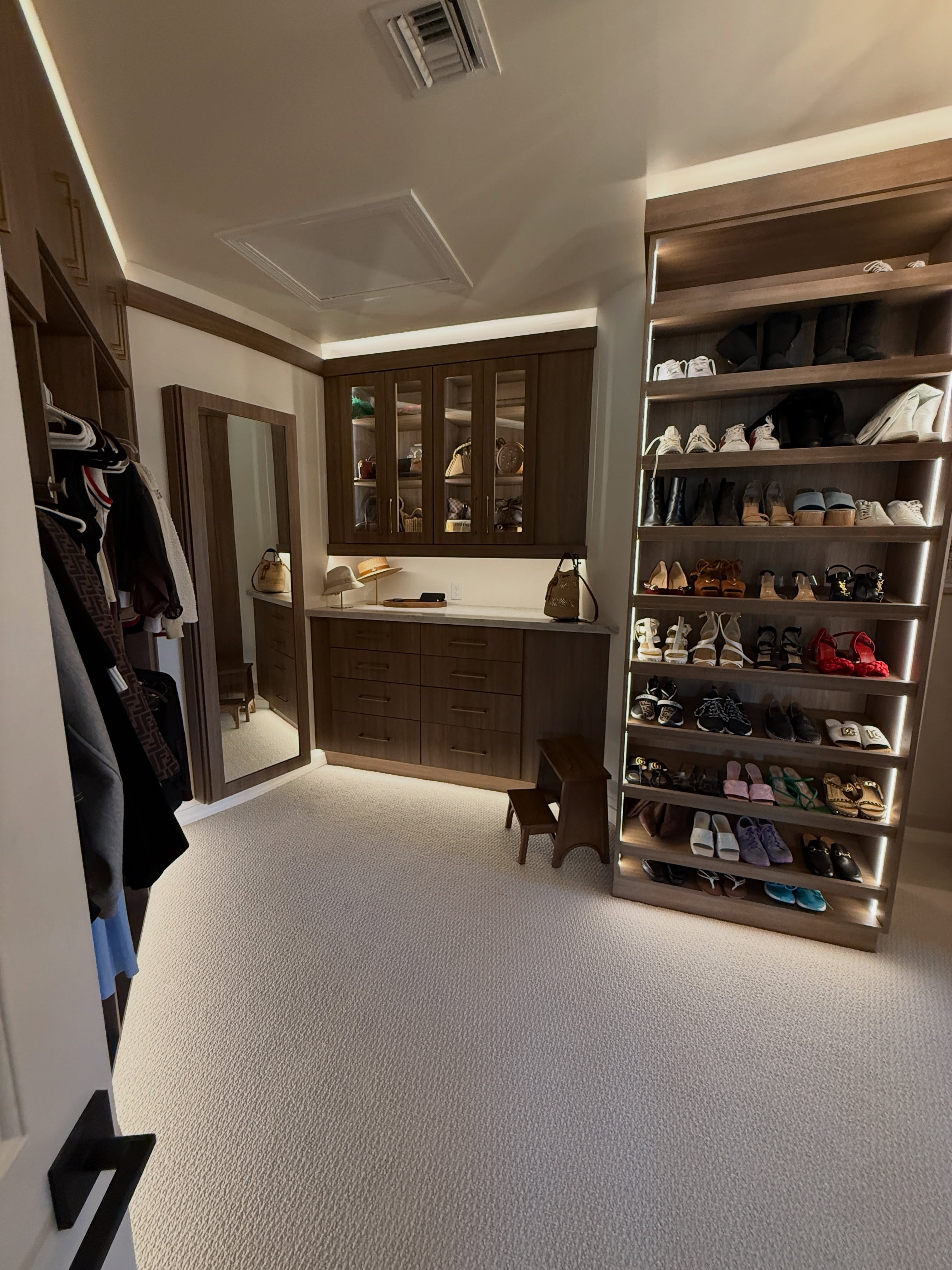 Completed Paradise Valley custom walk-in closet with dark wood built-in cabinets, glass-front cabinetry, illuminated shoe shelving, a mirror, counter, and carpet